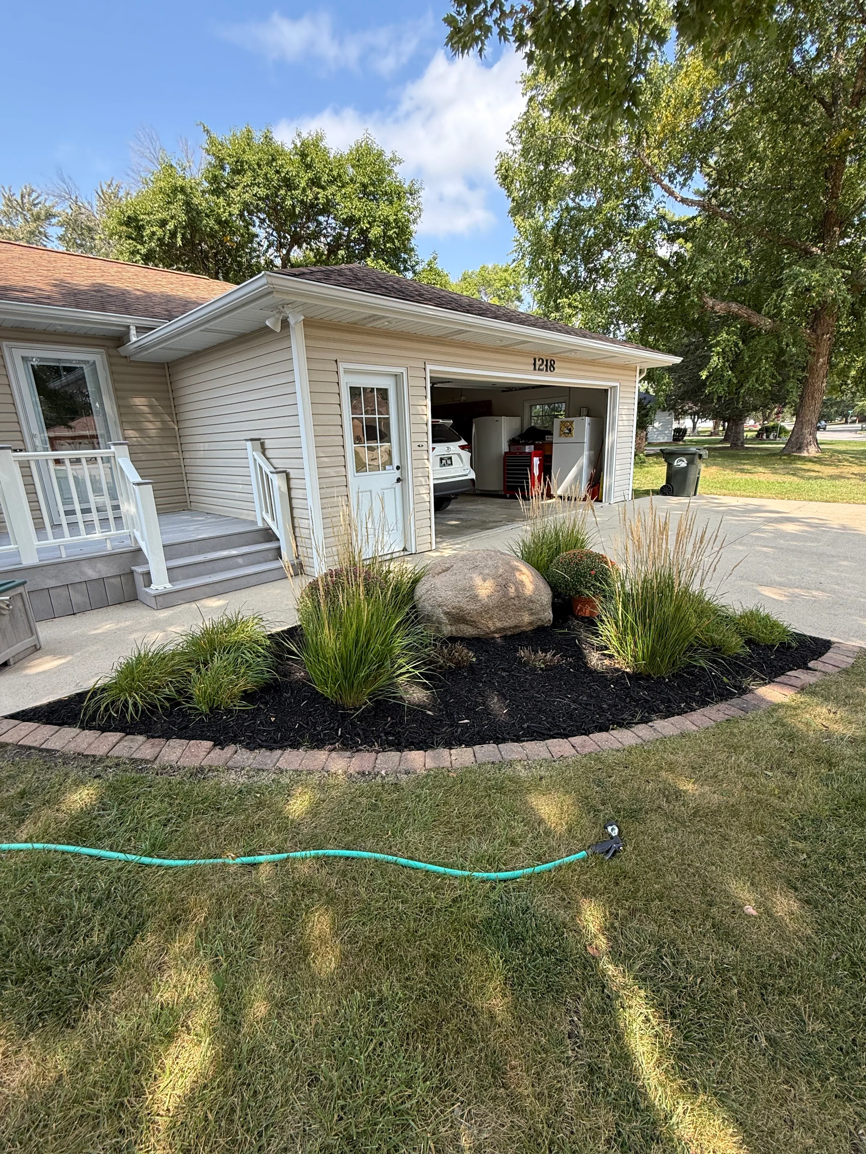 Front yard landscape bed with ornamental grasses, boulder, and black mulch bordered by brick edging